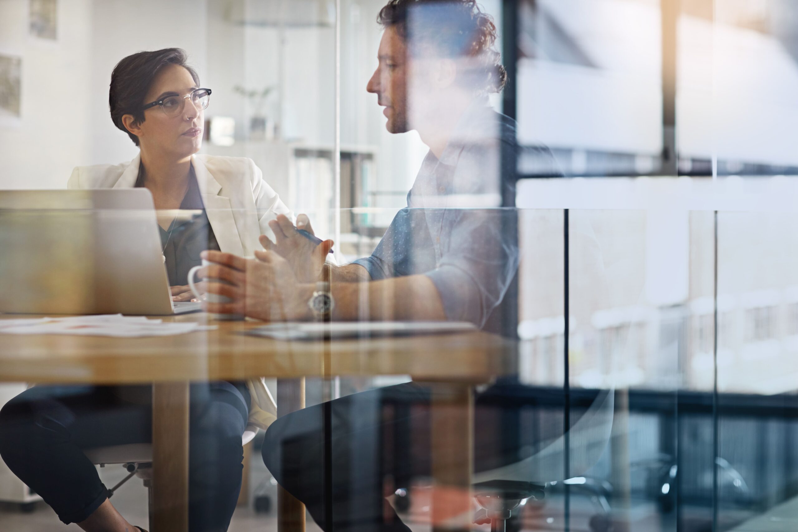 Two colleagues in a glass walled meeting room having a conversation.